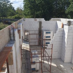 BudArtMagic Mykhailo Zagorodnyi - Wznoszenie ścian domu z białych bloczków silikatowych, widoczne rusztowanie, zbrojenie i palety z materiałem budowlanym na placu budowy w słoneczny dzień.