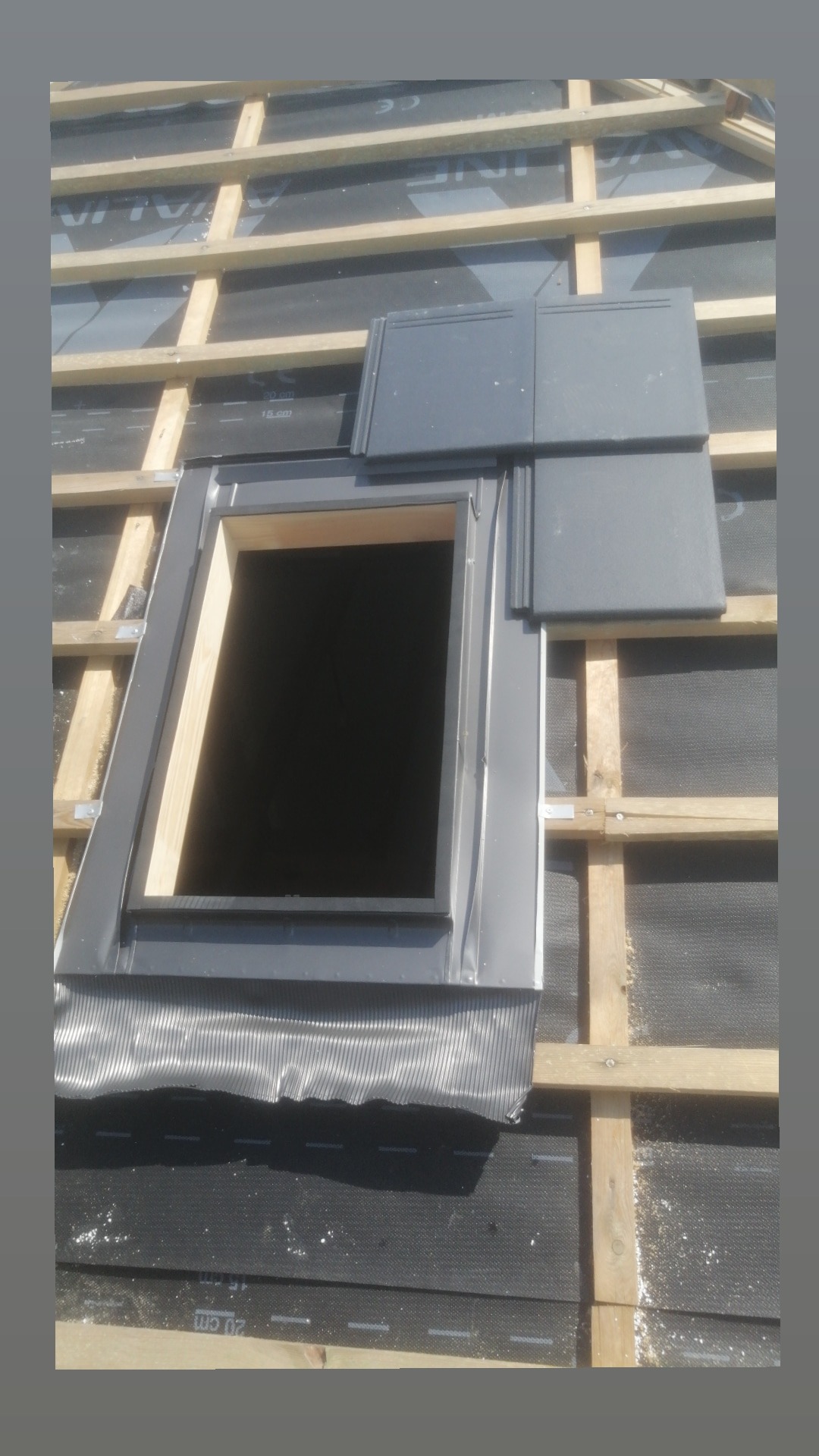 Montaż okna dachowego z obróbką blacharską i częściowo ułożoną dachówką ceramiczną w kolorze grafitowym, widoczne łaty drewniane i membrana dachowa.