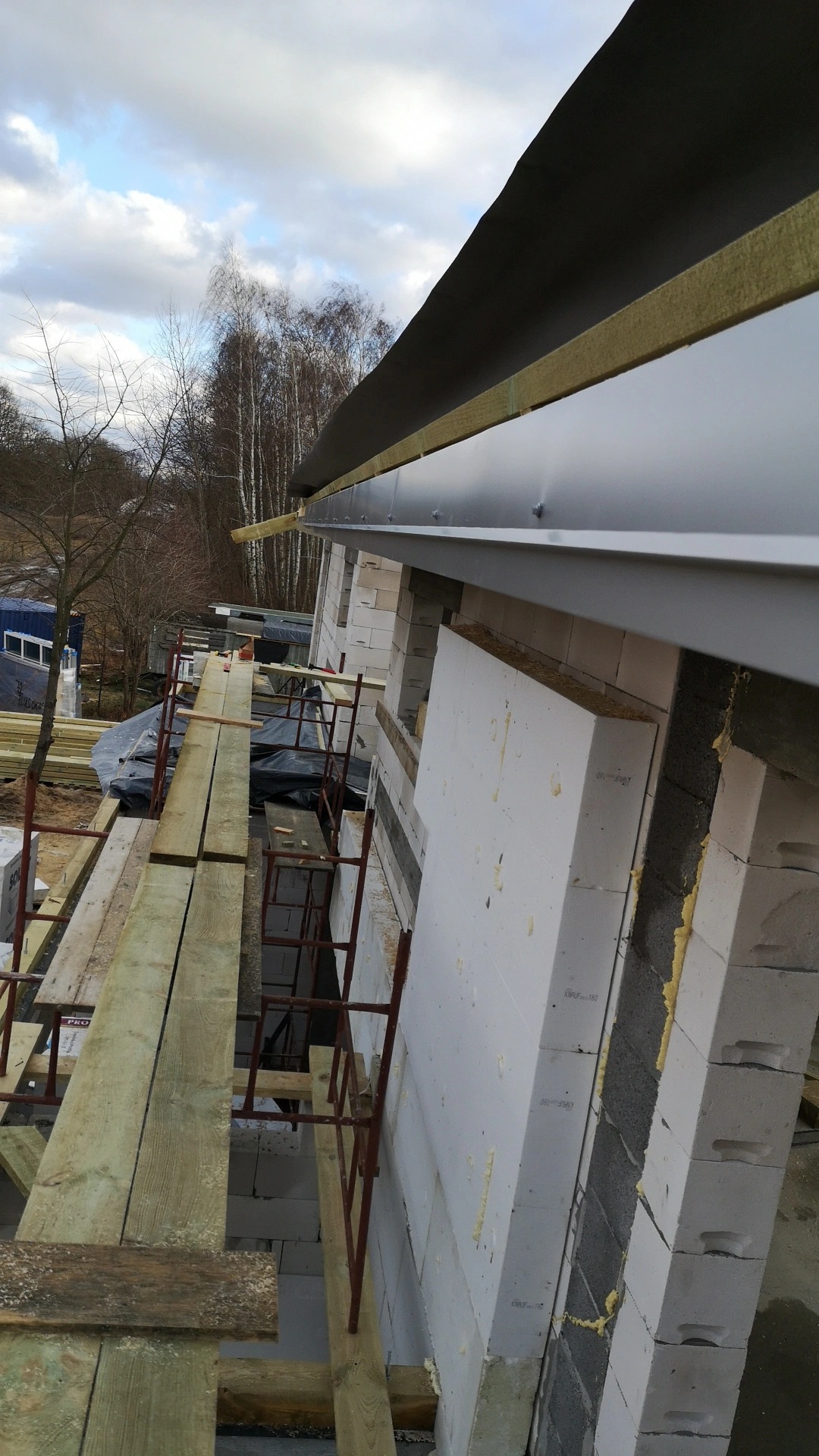 Budowa domu z białych bloczków, na rusztowaniu drewniane deski, widoczne ocieplenie styropianem i wełną mineralną na dachu, pochmurne niebo w tle.