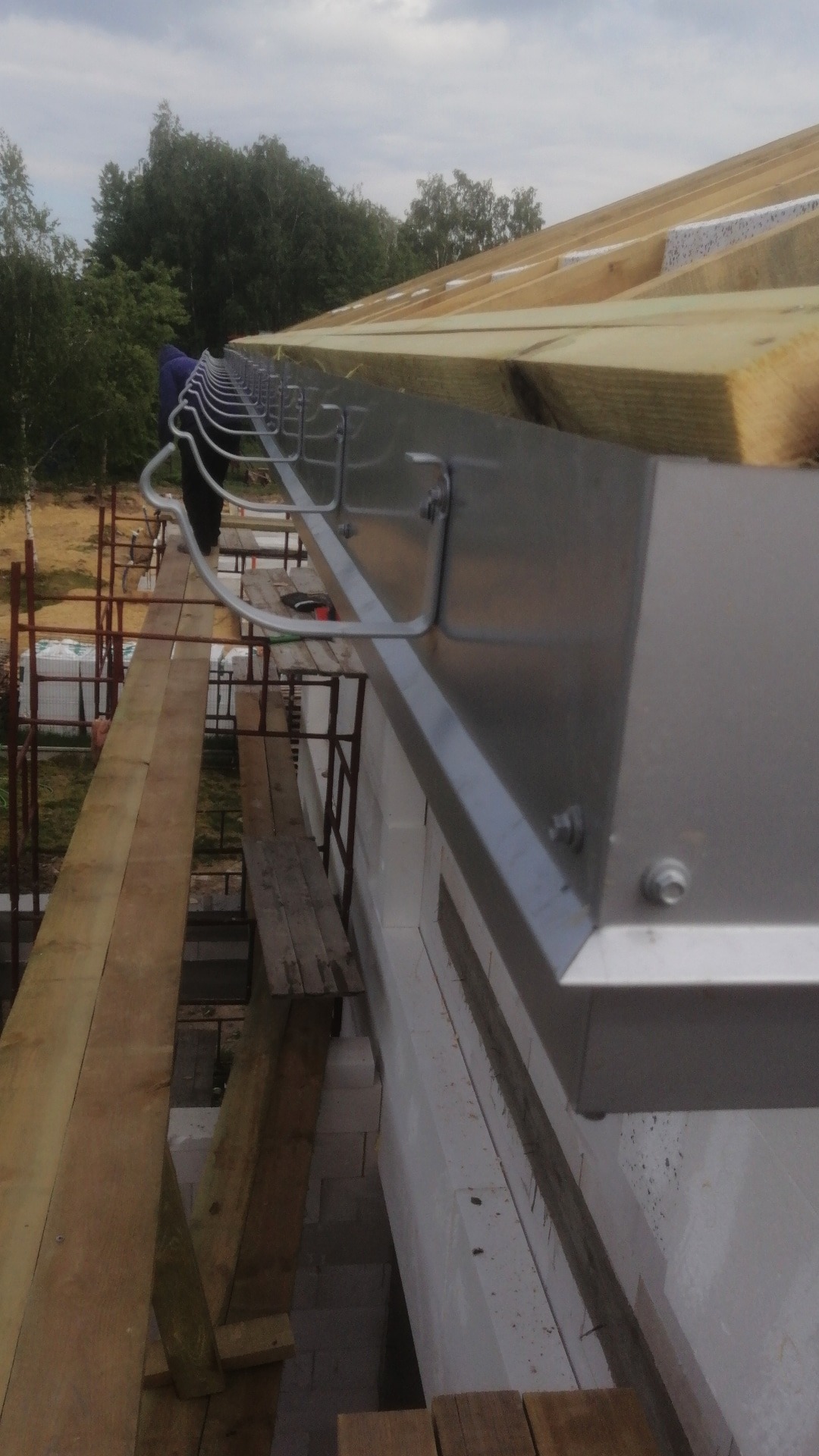 Montaż metalowej rynny na nowo budowanym domu z białego pustaka, widoczne krokwie z ociepleniem, rusztowanie i fragment sylwetki pracownika.