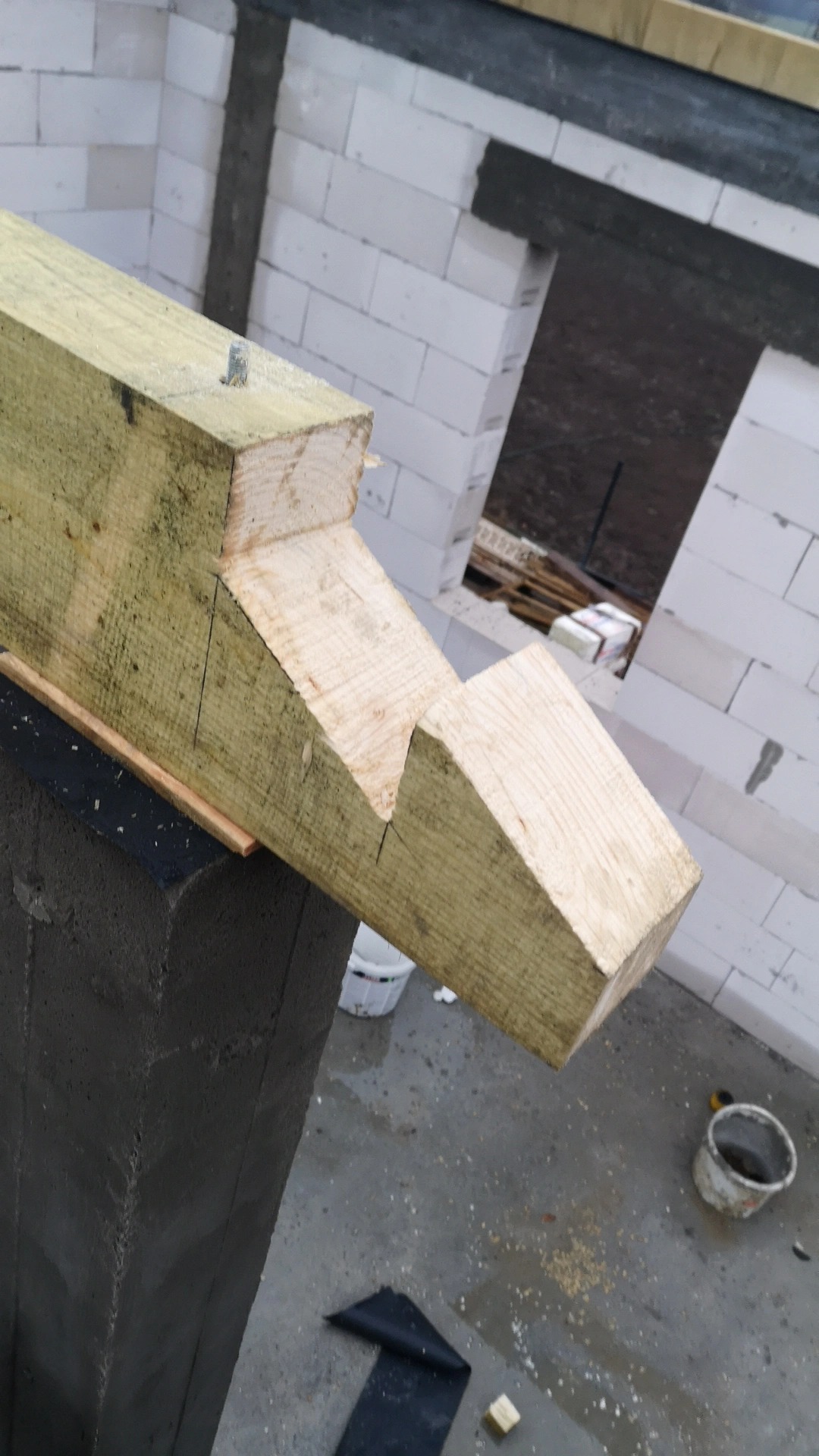 Fragment konstrukcji domu w trakcie budowy: drewniana belka z wycięciami oparta na szarej kolumnie, w tle białe bloczki ścian z otworem okiennym.