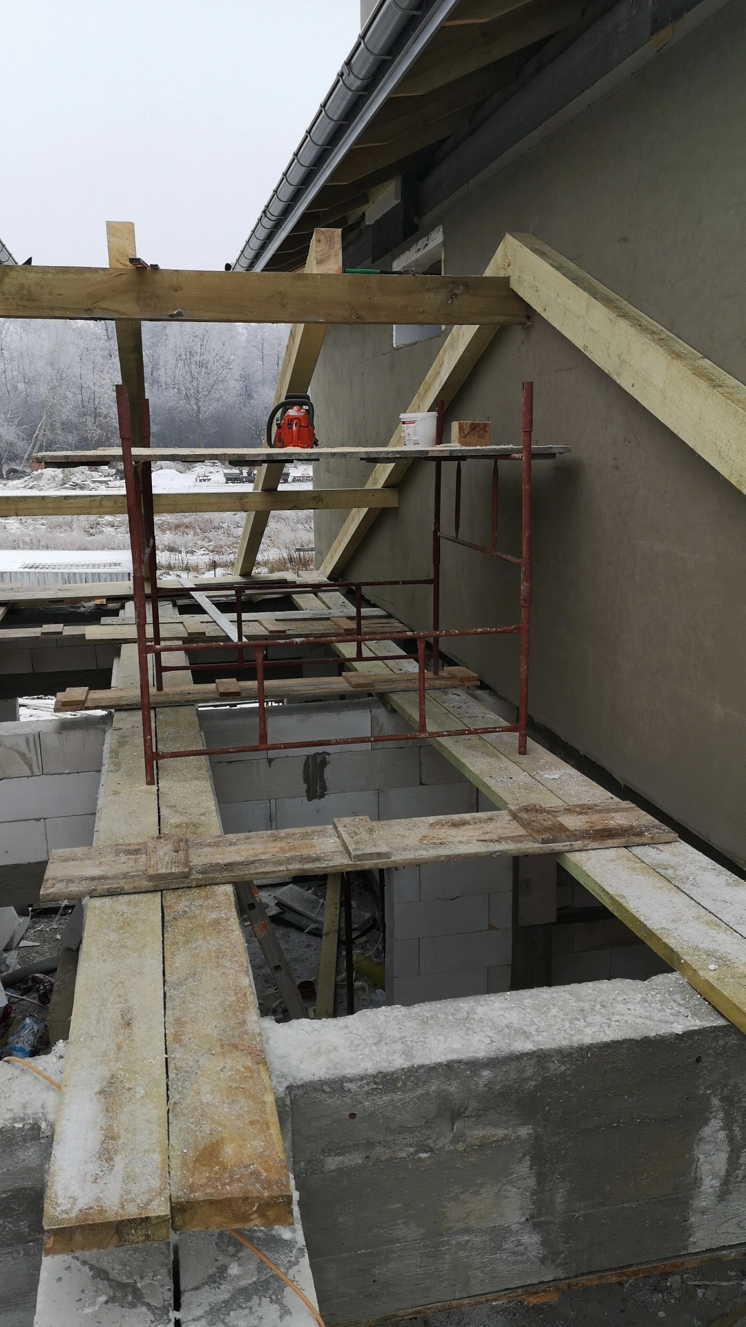 Wnętrze surowego, częściowo zadaszonego domu w trakcie budowy, widoczne białe bloczki, drewniane deski, metalowe rusztowanie i pomarańczowa piła spalinowa