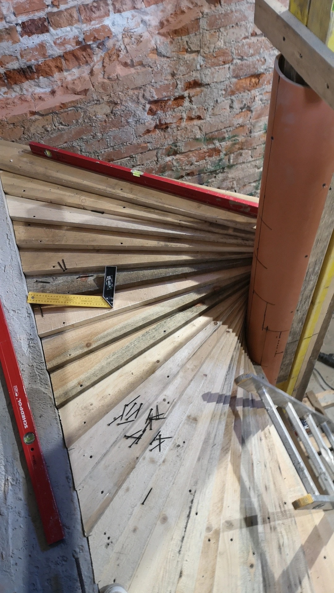 Wewnętrzna konstrukcja schodów spiralnych z drewnianych desek, widoczne poziomice, rura centralna i rozrzucone gwoździe na tle surowej, ceglanej ściany.