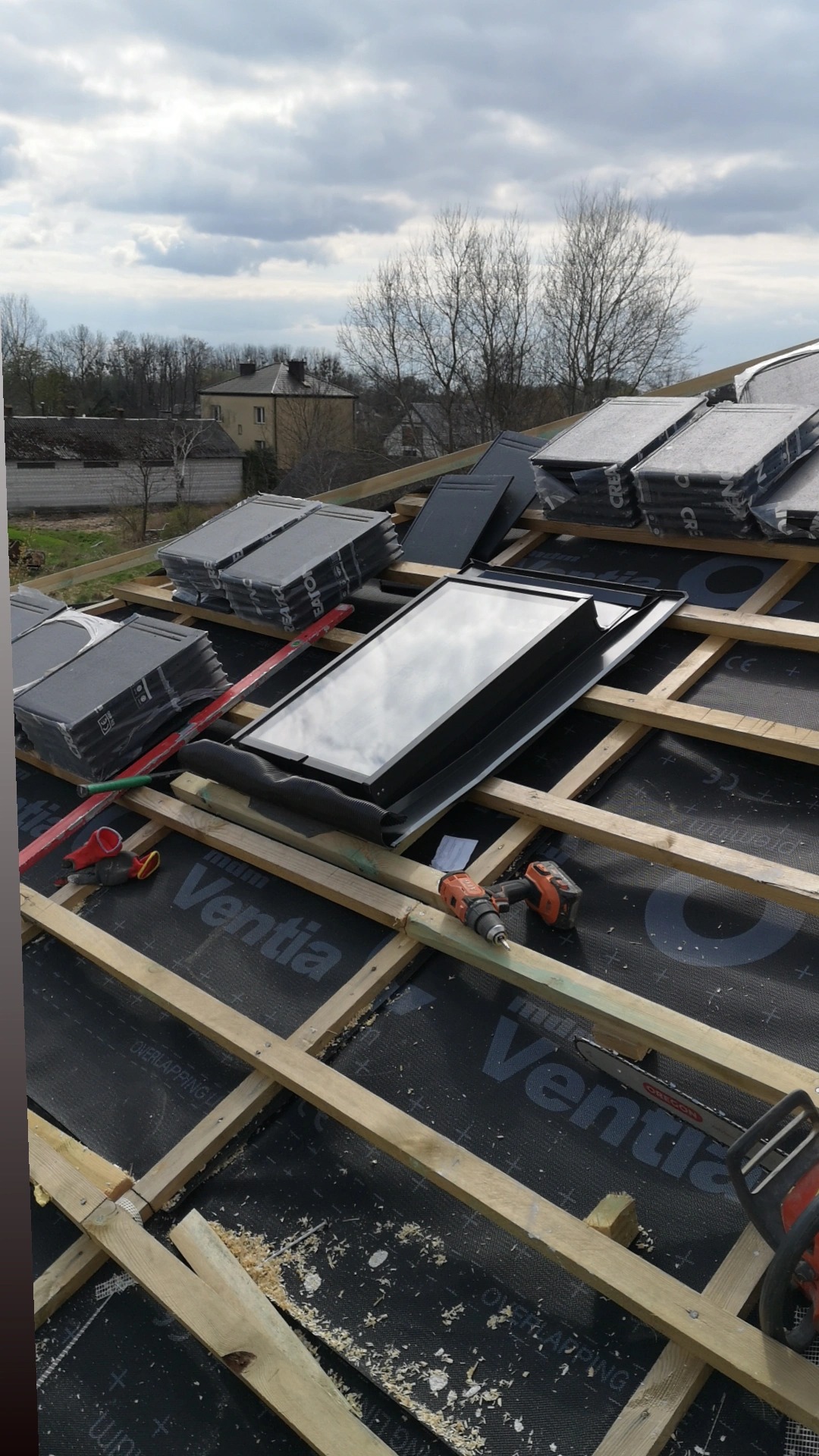 Montaż okna dachowego na konstrukcji drewnianej dachu pokrytej membraną, widoczne narzędzia, dachówki w opakowaniach i odległe budynki w tle.