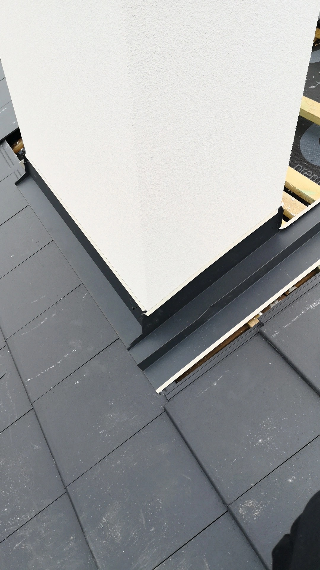 Szczegółowe ujęcie komina z szorstkim, białym wykończeniem, osadzonego w ciemnoszarym dachu z dachówki, z widoczną czarną obróbką blacharską i elementami konstrukcji drewnianej pod dachem.