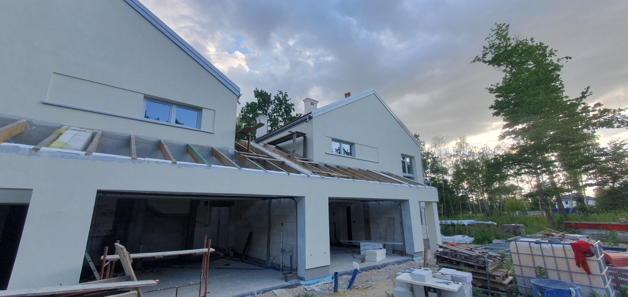 Dwa budynki w stanie surowym otwartym z widocznymi elementami konstrukcji dachu, jasna elewacja, okna wstawione, otoczenie placu budowy z materiałami budowlanymi.