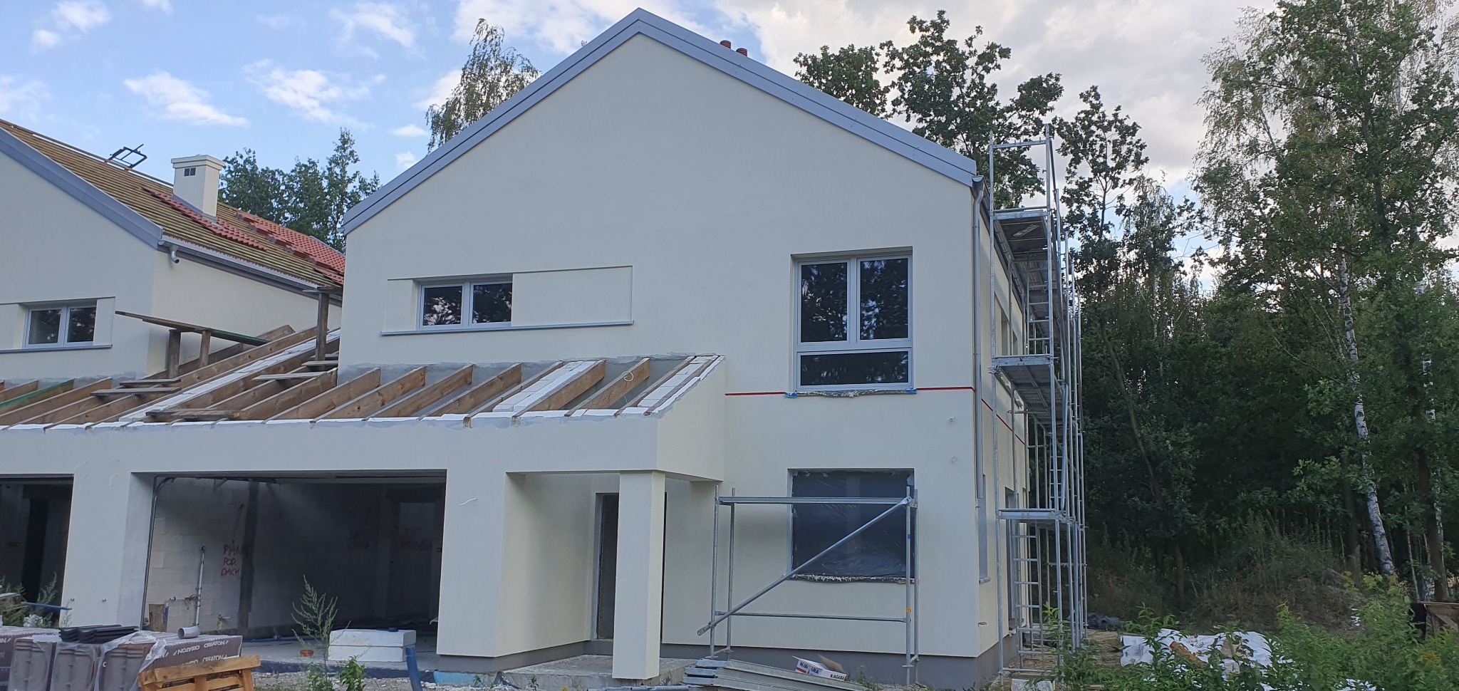 Nowo wybudowany dom jednorodzinny z garażem w stanie surowym zamkniętym, otynkowany na jasnożółto, z widocznymi rusztowaniami i elementami drewnianej konstrukcji dachu.