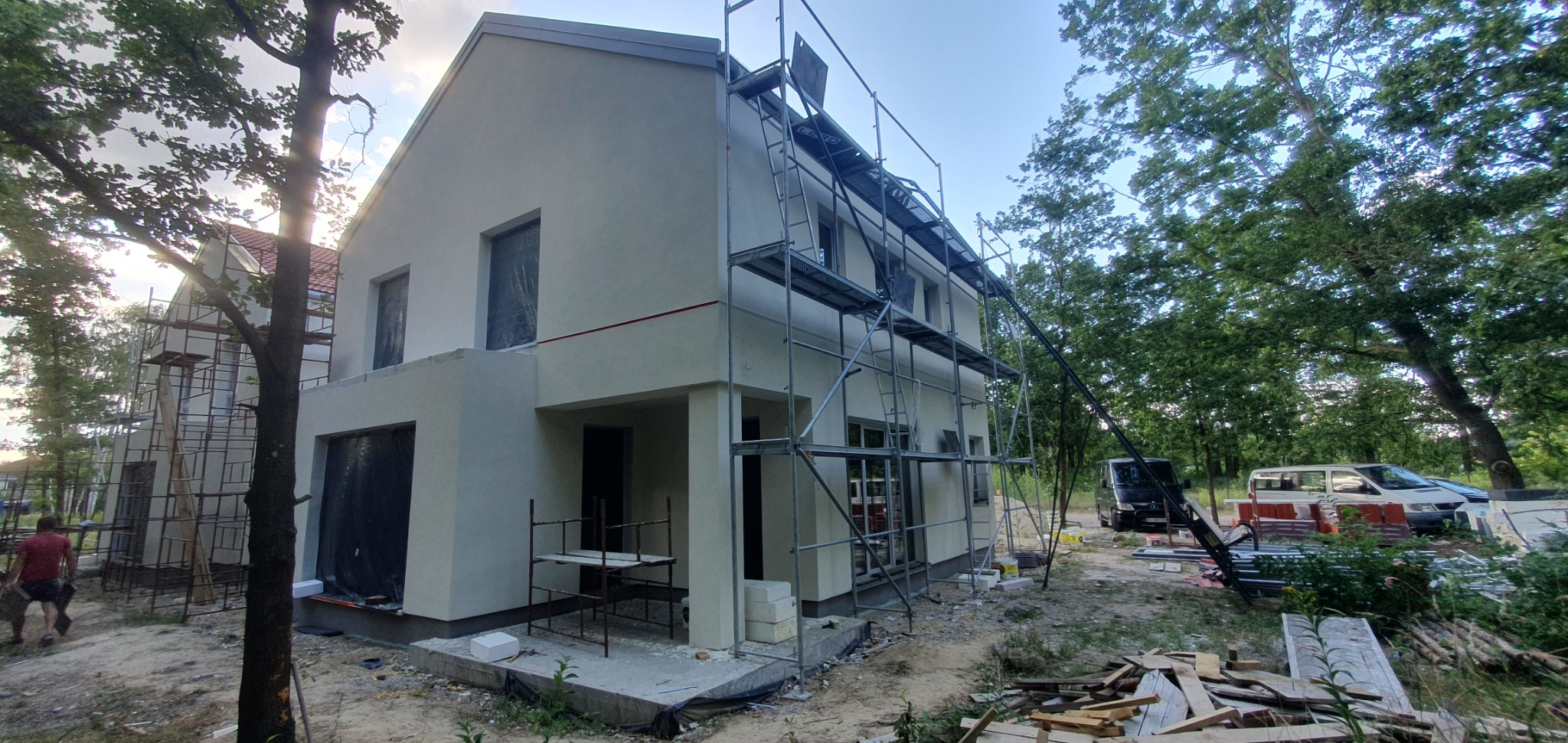 Dwukondygnacyjny dom w trakcie prac wykończeniowych elewacji, z widocznym rusztowaniem, zabezpieczonymi oknami i przygotowanym podłożem pod taras.