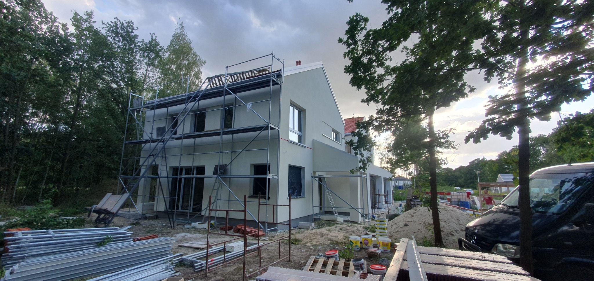 Dwupiętrowy dom murowany w trakcie ocieplania elewacji, z widocznymi rusztowaniami, niezabezpieczonymi oknami i stertą materiałów budowlanych na posesji, zarysowana sylwetka ciemnego auta w prawym...