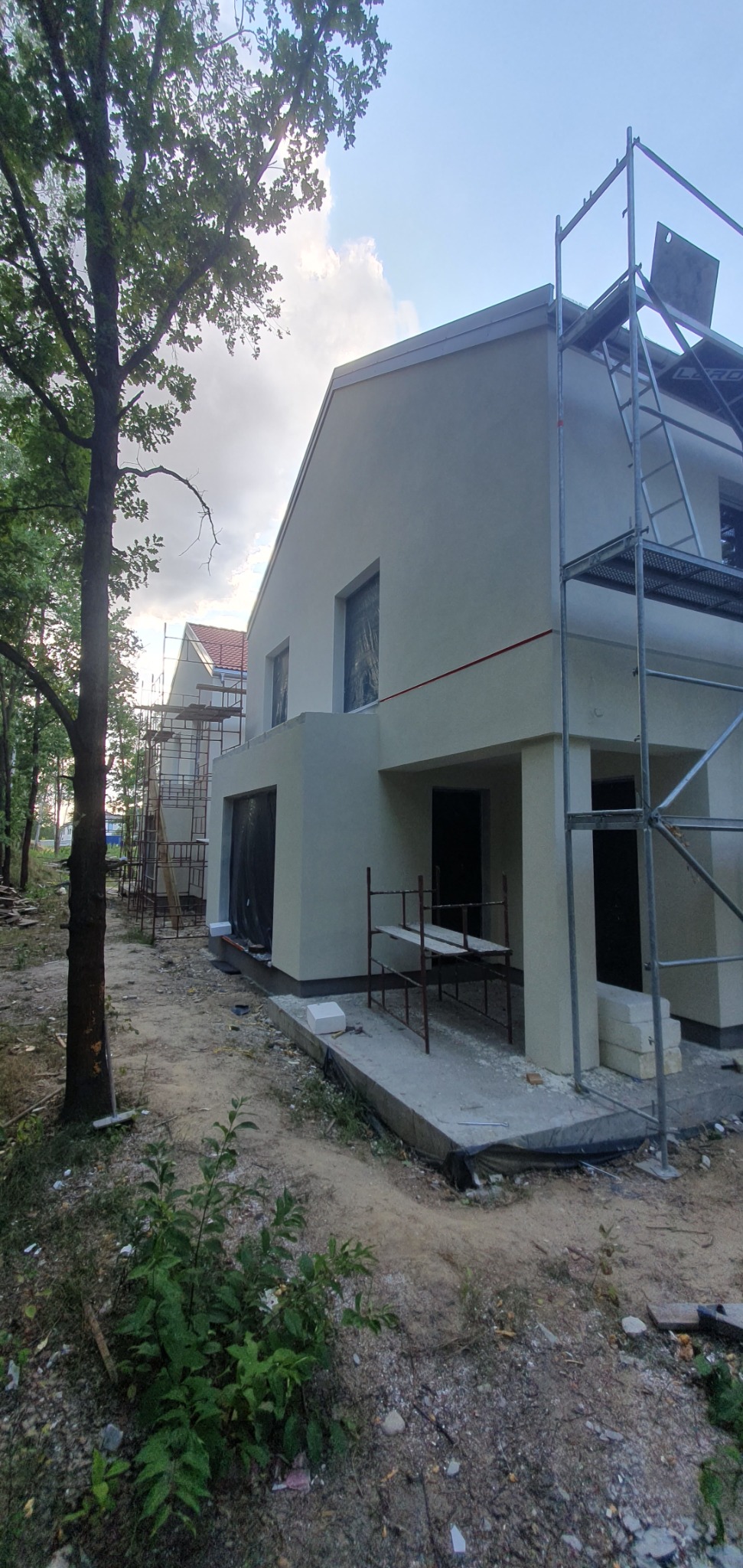Wznoszony dom murowany z szarym tynkiem, widoczne rusztowania i teren budowy w otoczeniu drzew.