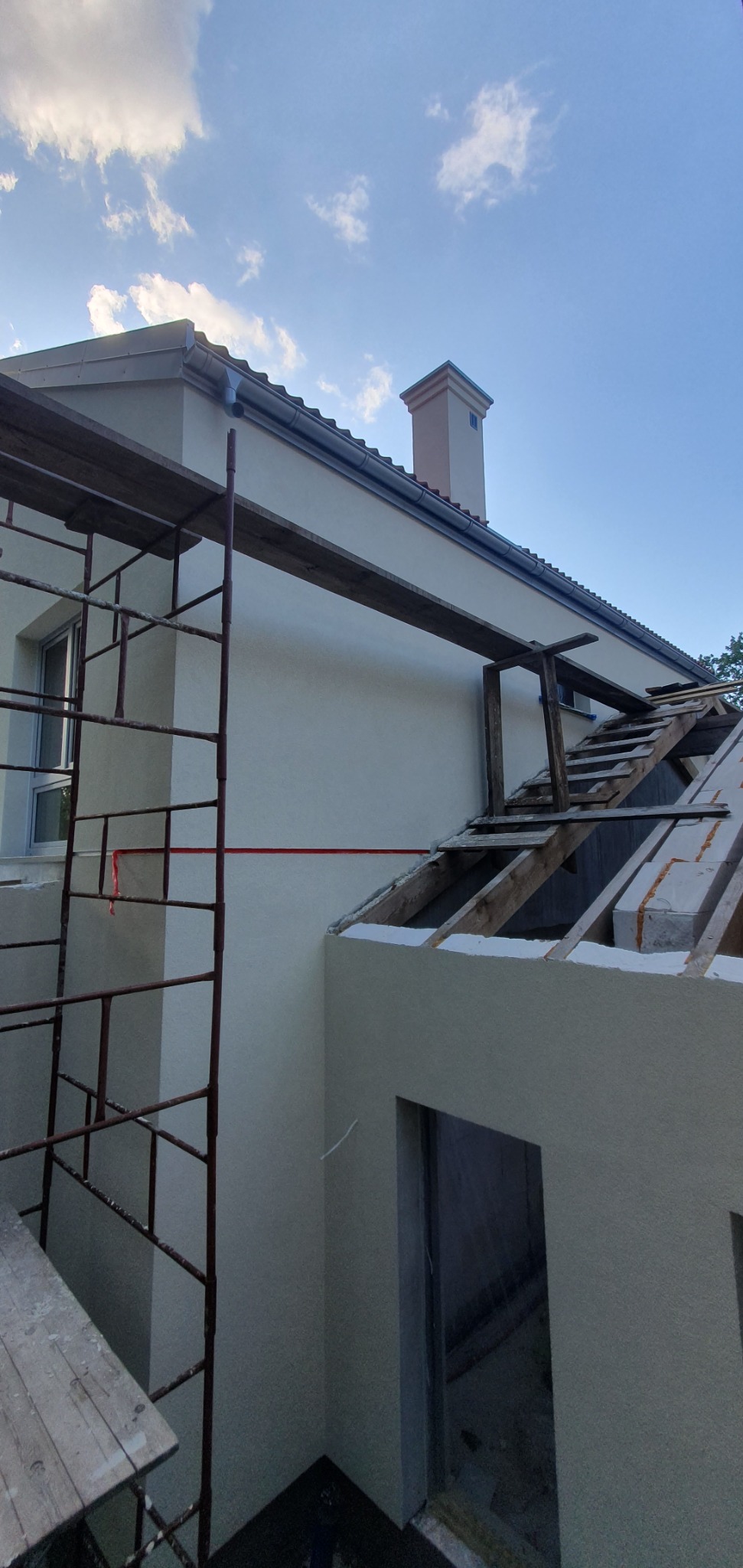 Budynek w trakcie prac budowlanych: murowana konstrukcja z rusztowaniem, widoczne elementy więźby dachowej i komin na tle błękitnego nieba.