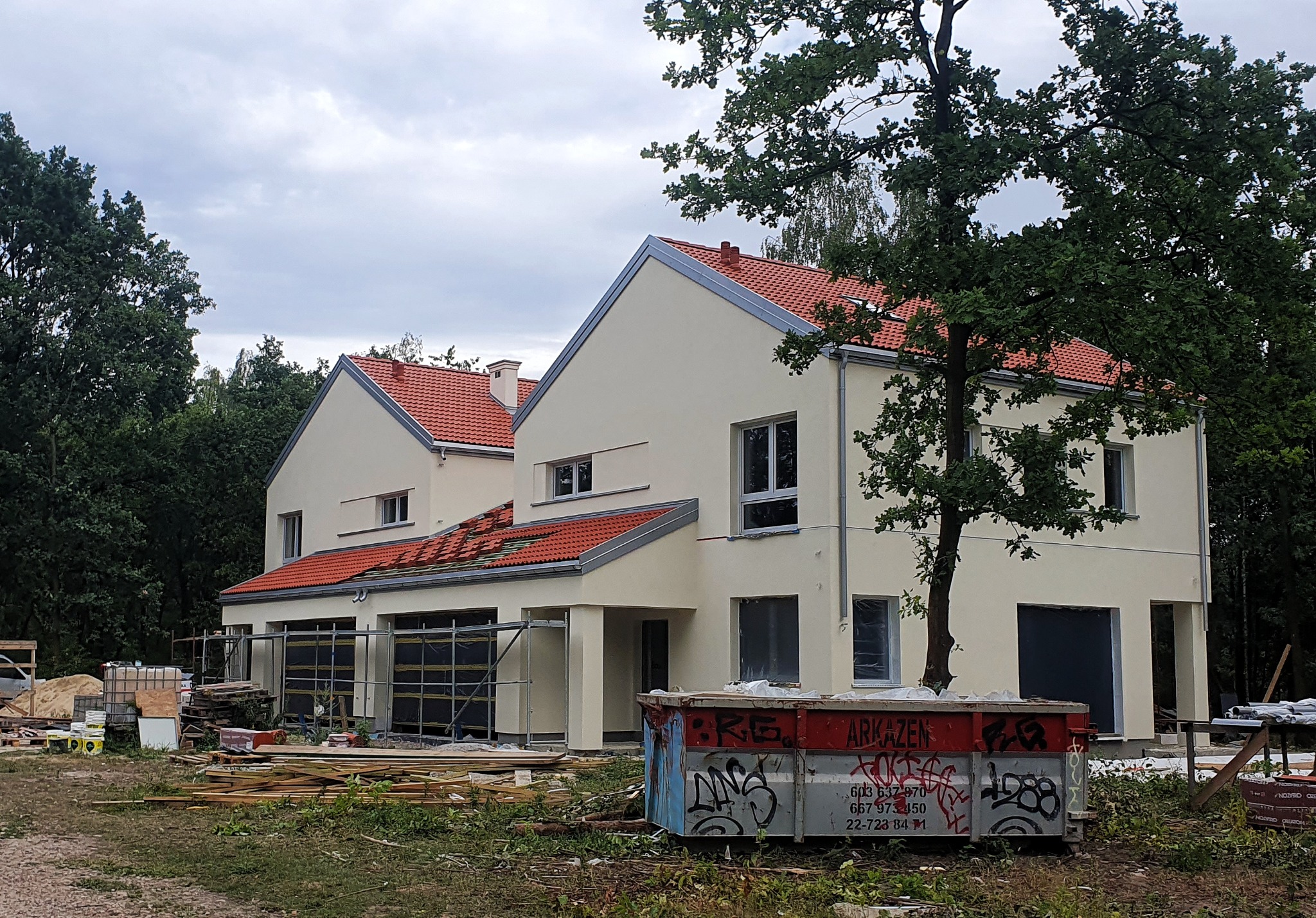 Nowoczesny, dwukondygnacyjny dom w trakcie budowy z czerwonym dachem, widoczny kontener na gruz z graffiti i materiały budowlane na działce.