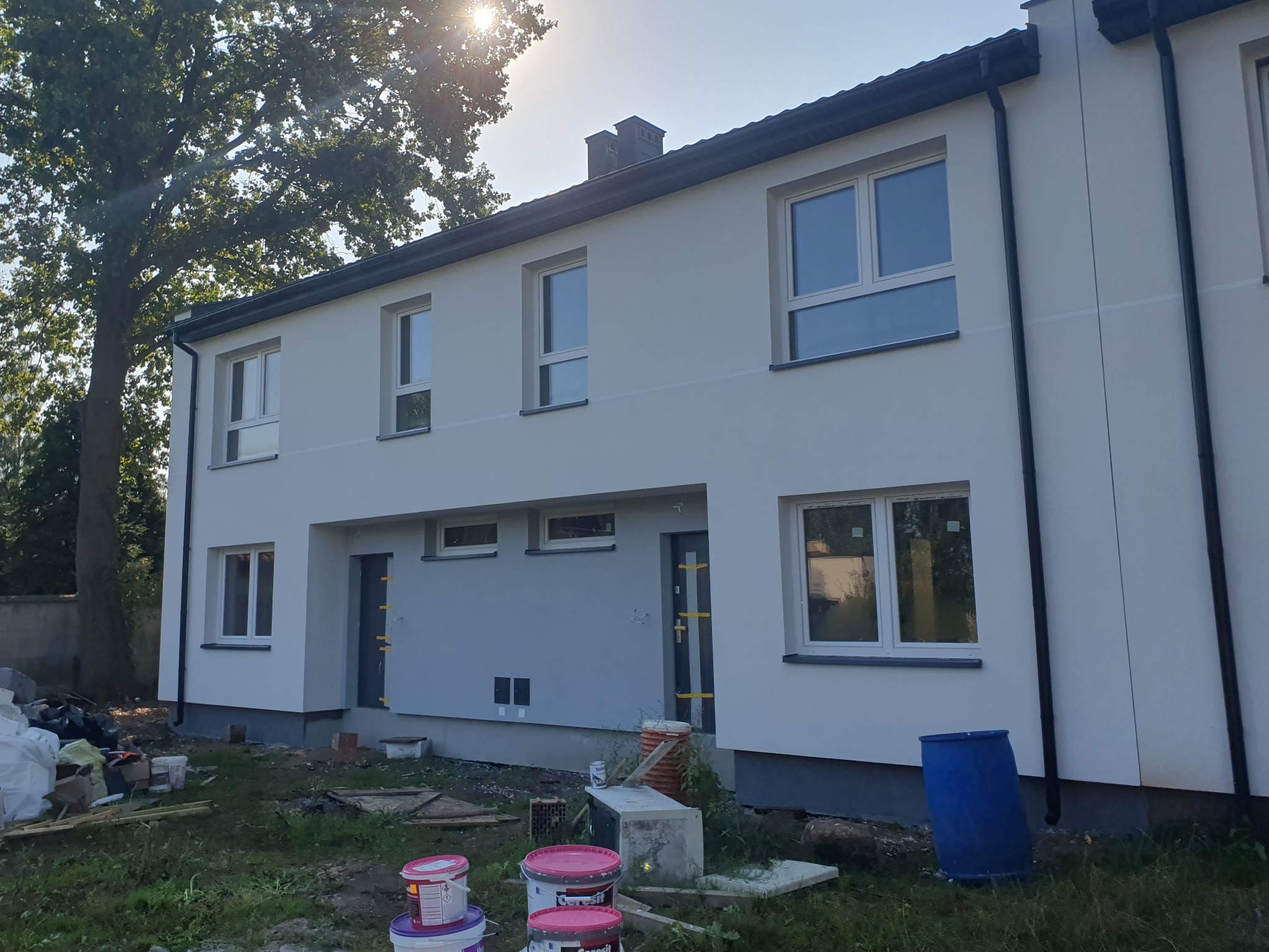 Bliźniak w trakcie prac wykończeniowych elewacji, widoczne puszki z farbą i niebieska beczka, na tle drzew i błękitnego nieba.