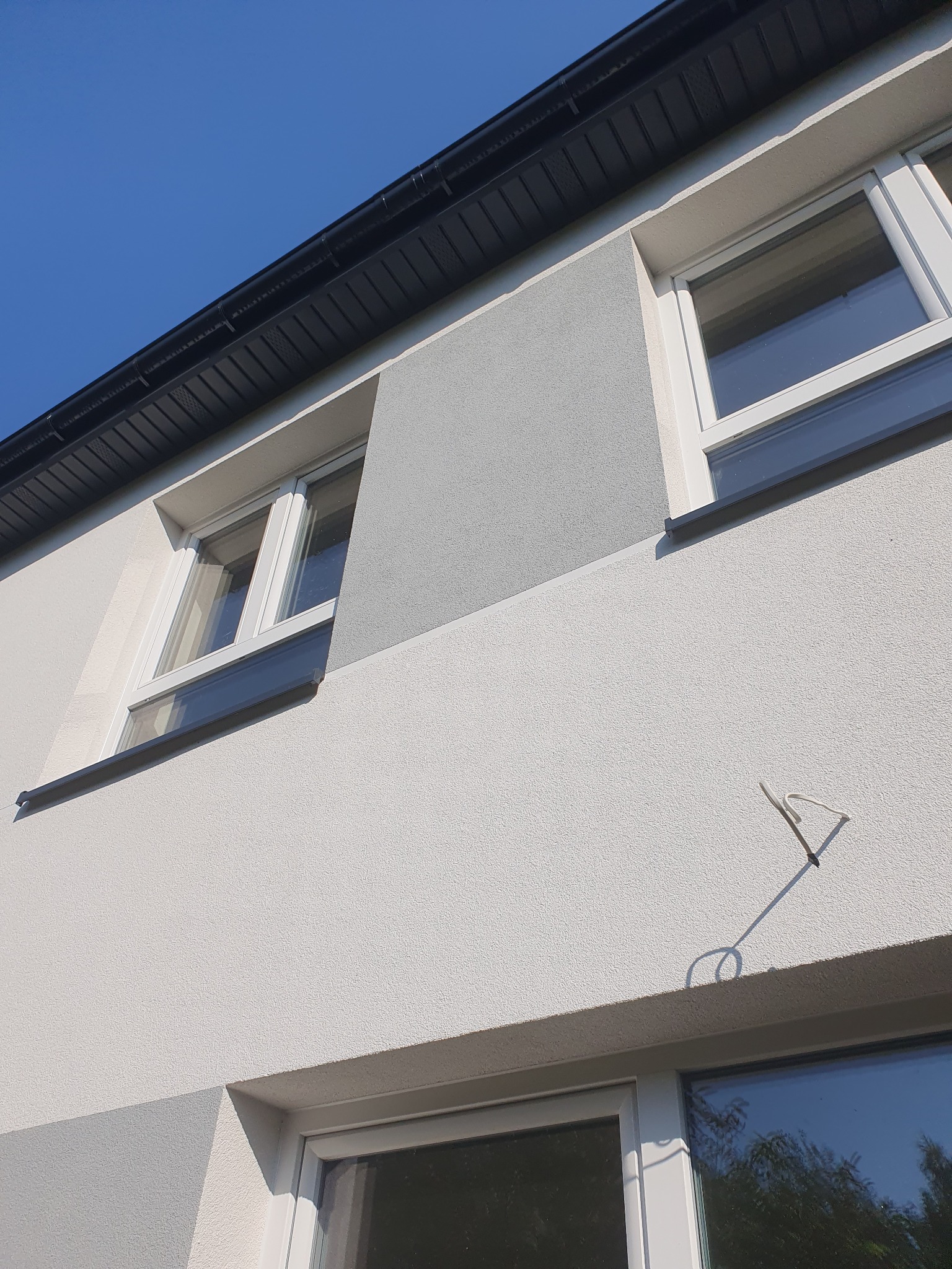 Fragment ocieplonej elewacji domu z dwoma oknami, widoczne różnice w odcieniach szarości tynku oraz czarny orynnowanie pod błękitnym niebem.