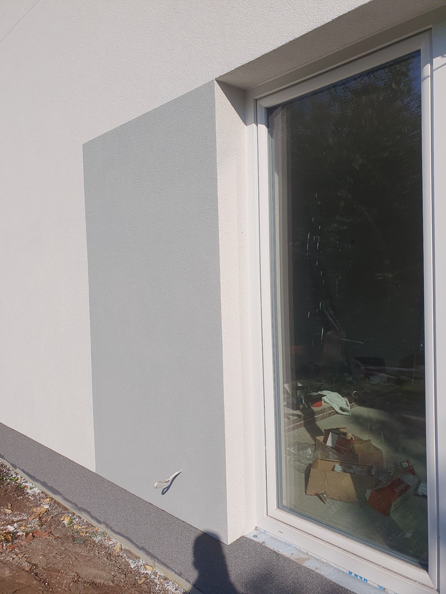 Fragment elewacji budynku z oknem, widoczna struktura tynku w odcieniach bieli i szarości, oraz nieporządek w pomieszczeniu widoczny przez szybę.