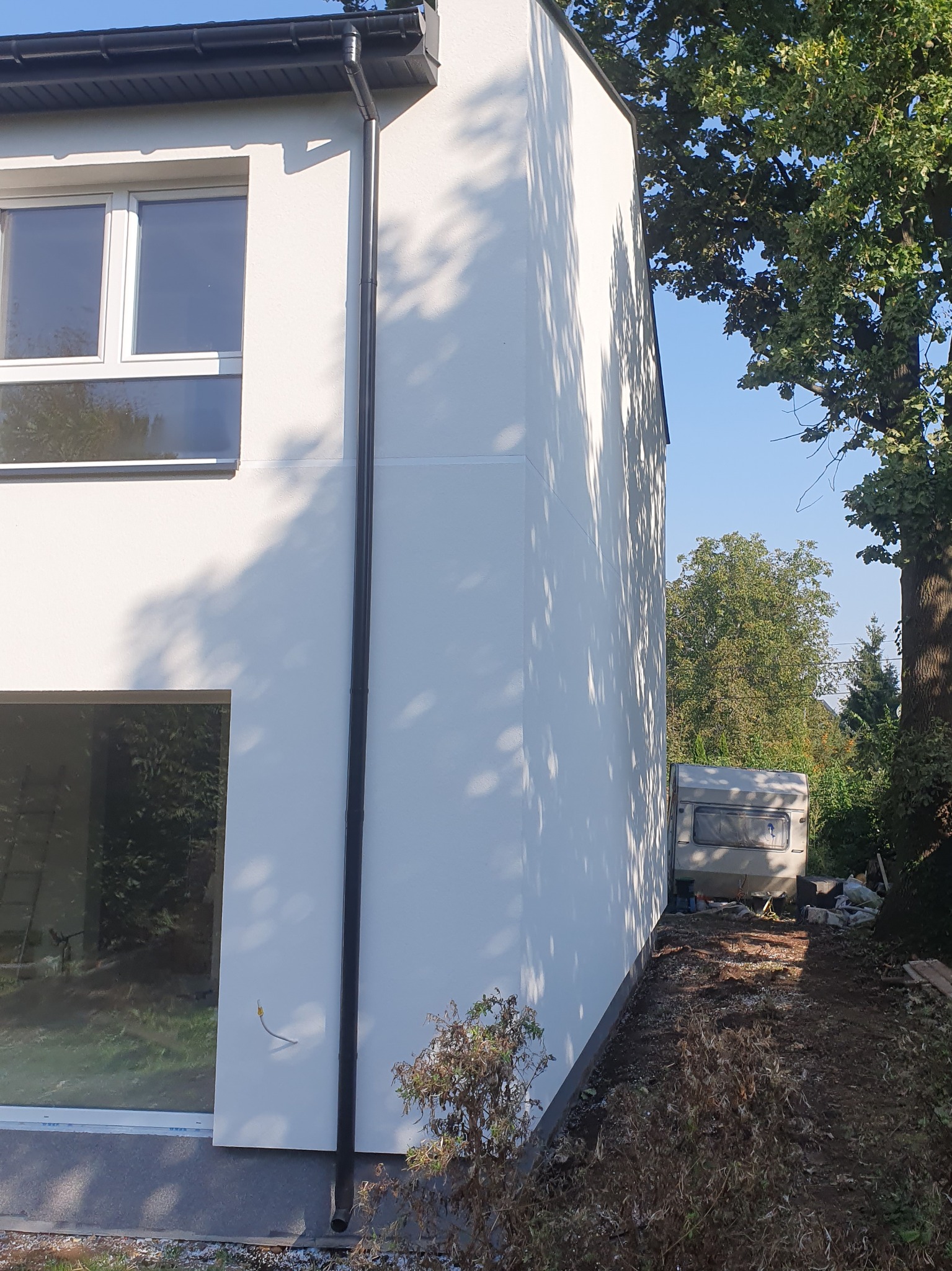Fragment bocznej ściany nowo wybudowanego, otynkowanego domu z oknami, rynną spustową i widocznym przyczepą kempingową w tle.