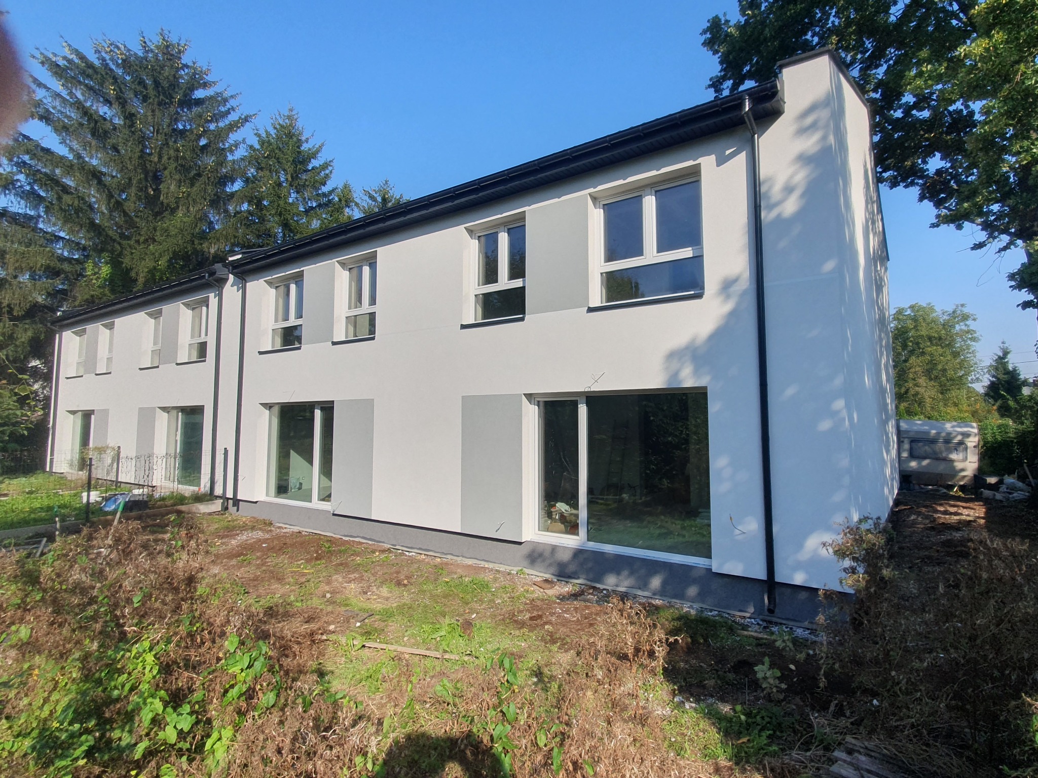 Nowoczesny, szeregowy budynek mieszkalny z białymi ścianami i szarymi akcentami, duże okna, otoczony zielenią i elementami krajobrazu w trakcie zagospodarowywania.