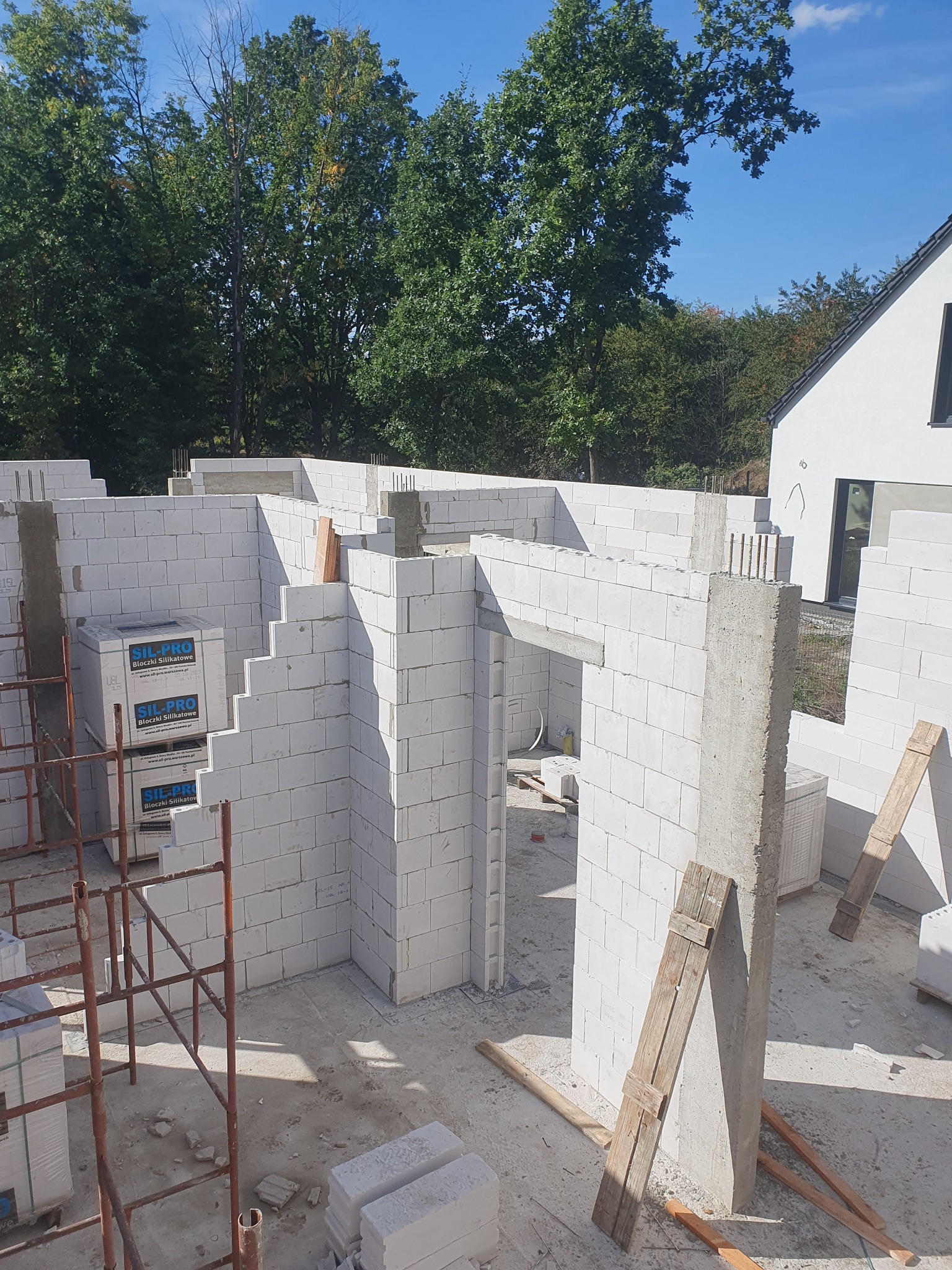 Wznoszenie ścian domu z białych bloczków silikatowych SIL-PRO, widoczne stalowe rusztowanie, drewniane podpory i fragment budynku w tle.