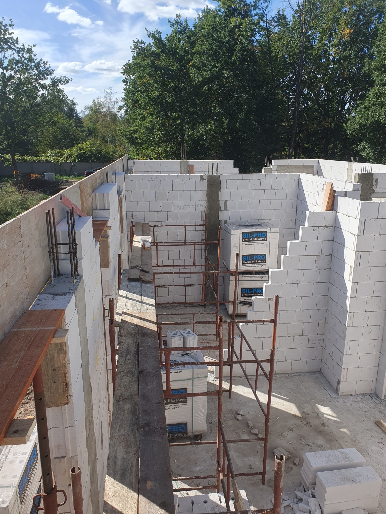 Wznoszenie ścian domu z białych bloczków silikatowych, widoczne rusztowanie, zbrojenie i palety z materiałem budowlanym na placu budowy w słoneczny dzień.