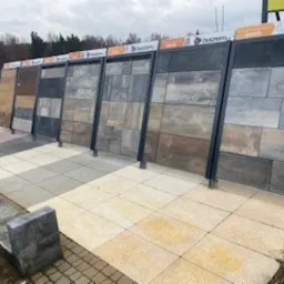 Ekspozycja różnorodnych wzorów i kolorów nawierzchni ułożonych na placu, z widocznymi tabliczkami informacyjnymi i ławką z betonu architektonicznego w tle.