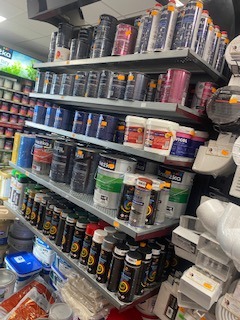 Półki w sklepie z materiałami budowlanymi, wypełnione różnorodnymi farbami w puszkach i butelkach, widoczne etykiety producentów, oświetlenie górne, perspektywa z dołu.