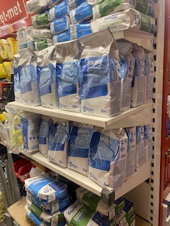 Półki w sklepie z materiałami budowlanymi, wypełnione workami z zaprawą murarską i klejami w niebiesko-białych opakowaniach. Widoczne etykiety z wagą 5 kg i logo producenta.