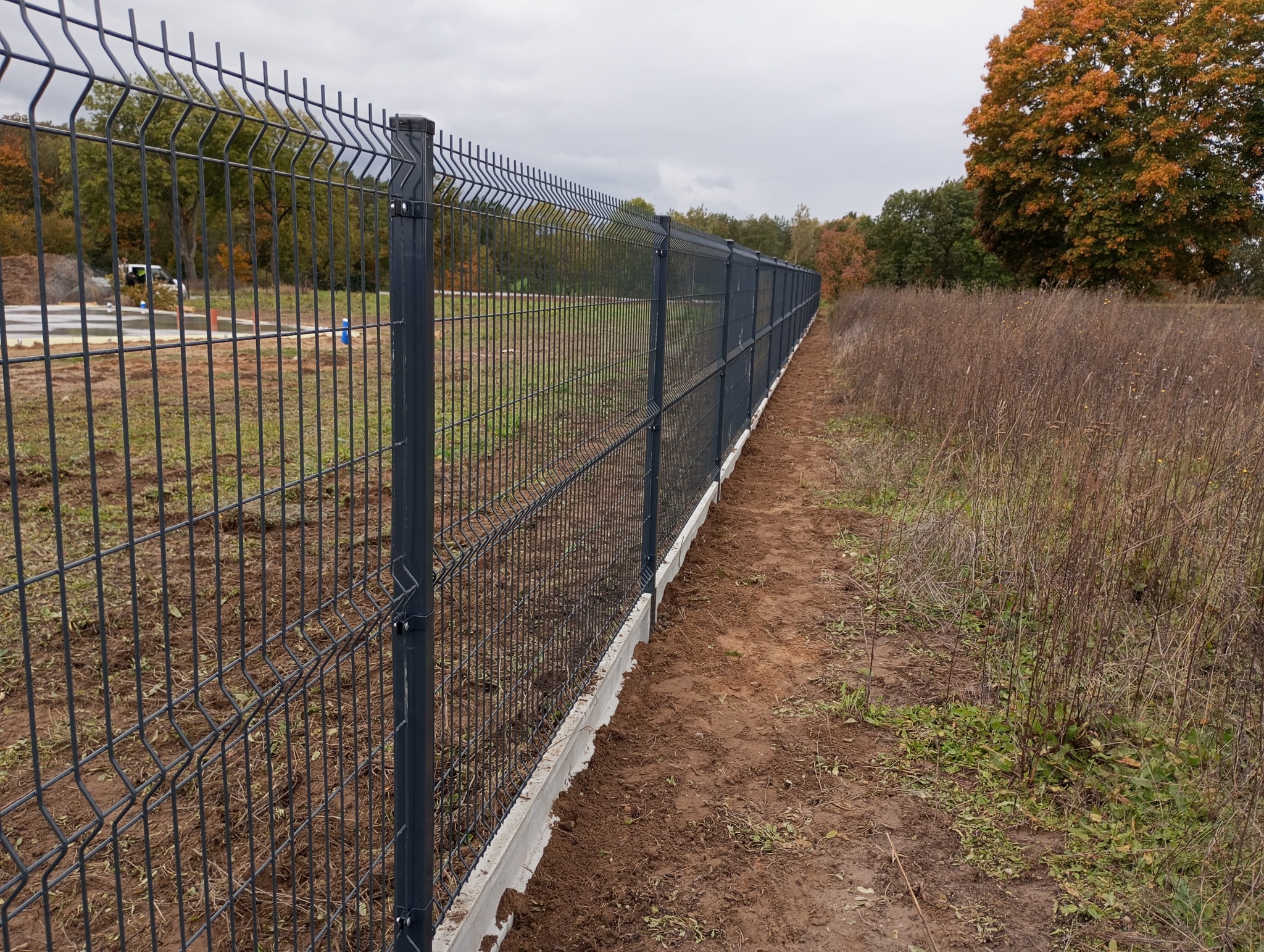 Długi, szary panel ogrodzeniowy z betonową podmurówką, ciągnący się wzdłuż polnej drogi, z drzewami w tle i pochmurnym niebem. Widok z perspektywy.