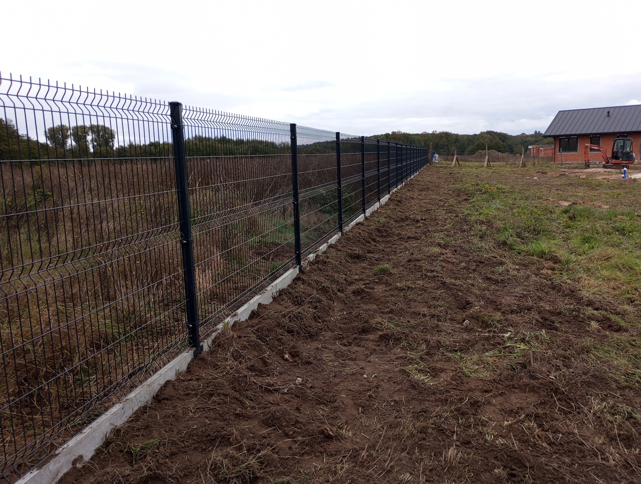 Długi, czarny panel ogrodzeniowy z podmurówką betonową, rozciągający się wzdłuż nierównego terenu z widocznym domem w budowie w tle, pochmurne niebo.