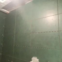 Ściana w łazience wyłożona zielonymi, matowymi płytkami ceramicznymi, widoczne krzyżyki dystansowe pomiędzy płytkami, fragment instalacji wentylacyjnej pod sufitem.