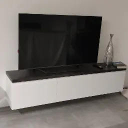 Nowoczesny salon z białą szafką RTV z ciemnym blatem, na której stoi telewizor, szklana dekoracja i książka. Obok stoi czarny hoker na srebrnej nodze. W tle widoczne drzwi balkonowe.