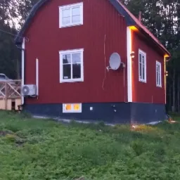 Czerwony dom z białymi ramami okiennymi i anteną satelitarną na ścianie, otoczony zielenią.