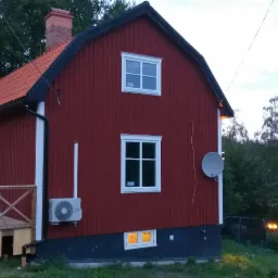 Czerwony drewniany dom z białymi ramami okiennymi i dachem z dachówki, widok z zewnątrz, z oświetlonym gankiem i anteną satelitarną na ścianie bocznej.