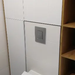 Nowoczesna toaleta podwieszana z białymi płytkami ściennymi z poziomymi żłobieniami, srebrnym przyciskiem spłukującym i drewnianym regałem z półkami po prawej stronie.