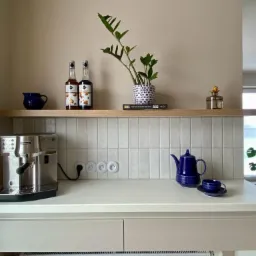 Jasna kuchnia z białymi płytkami w pionowy wzór, srebrnym ekspresem do kawy, niebieskim serwisem do herbaty i drewnianą półką z dekoracjami.