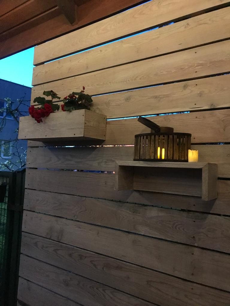 Drewniana ściana z poziomych desek z zamontowanymi półkami: skrzynka z czerwonymi kwiatami i półka z dekoracyjnym lampionem oraz świecą.