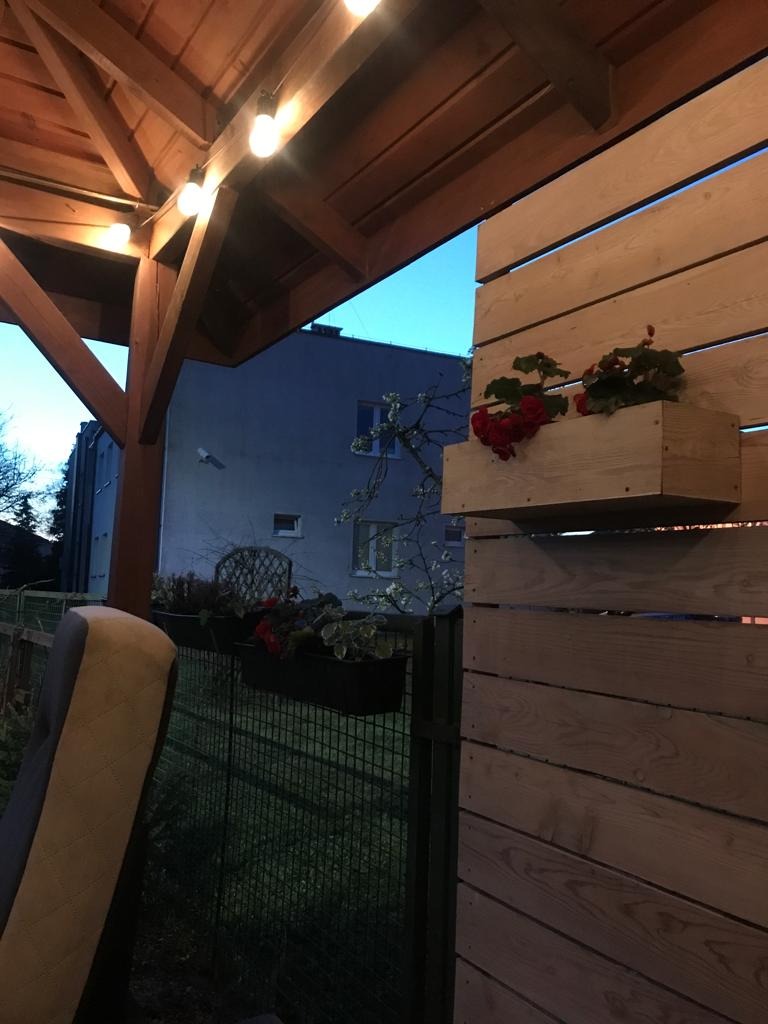 Drewniana altana z oświetleniem i skrzynkami z czerwonymi kwiatami na drewnianym panelu ogrodzenia, widok na budynek w tle.