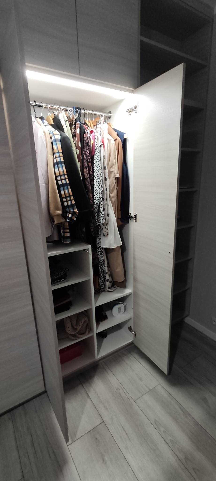 Otwarta szafa wnękowa z jasnego drewna, oświetlona od wewnątrz, z ubraniami na wieszakach i półkami z różnymi przedmiotami, widok z dołu.