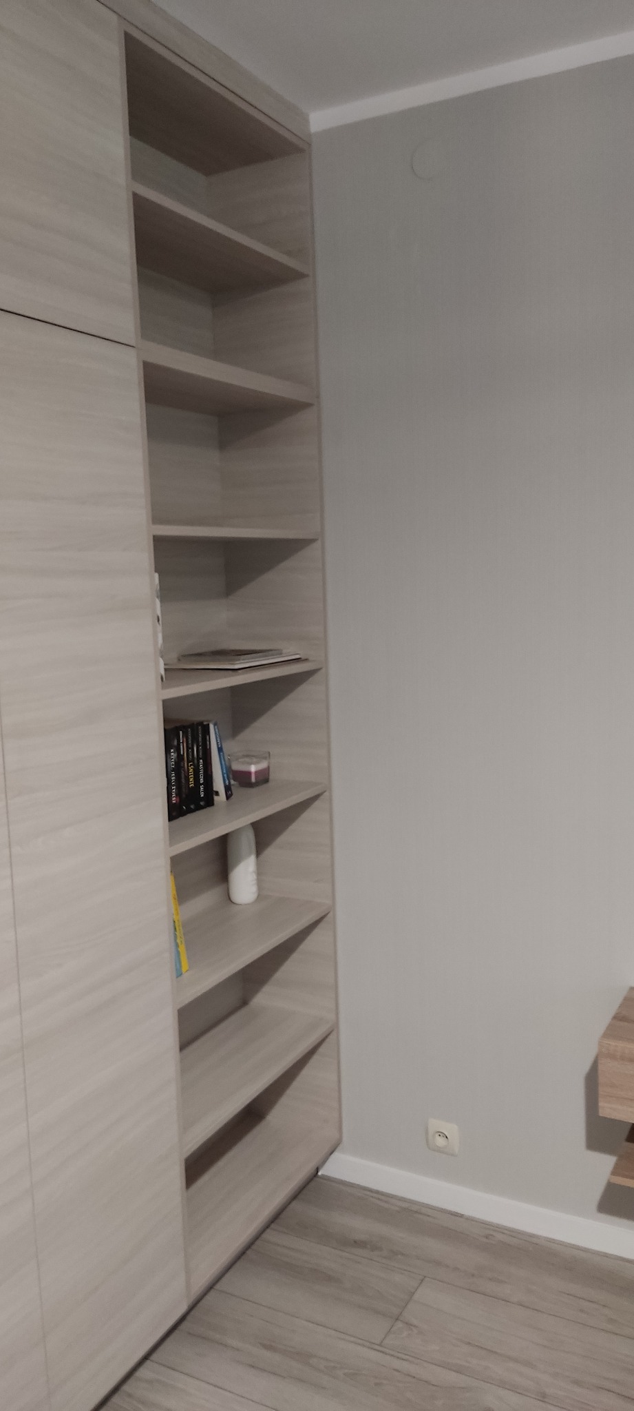 Jasna szafa wnękowa z półkami, na których ustawione są książki, świeca i dekoracyjny wazonik. Widoczna struktura drewna na frontach szafy i półkach. Na ścianie gniazdko elektryczne.