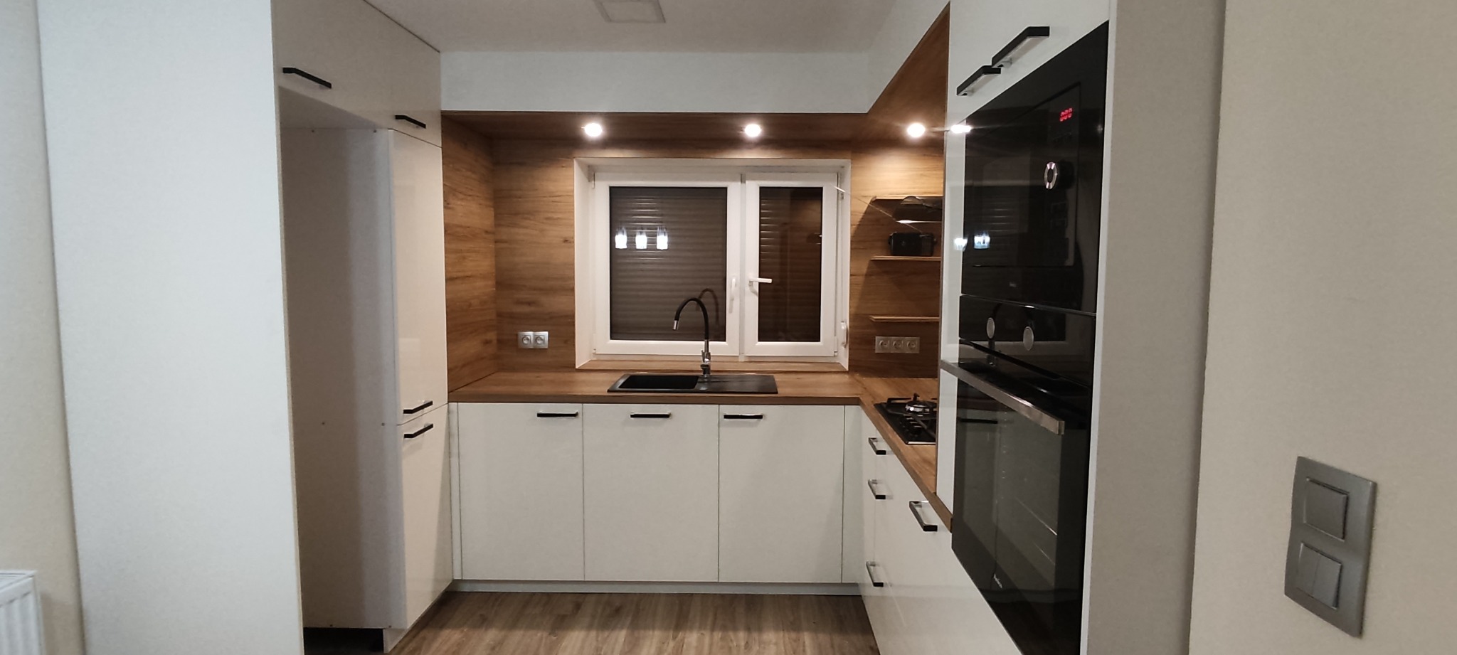 Nowoczesna kuchnia z białymi frontami, drewnianym blatem i okładziną ścienną, czarną armaturą i piekarnikiem w zabudowie, oświetlona punktowymi lampami.