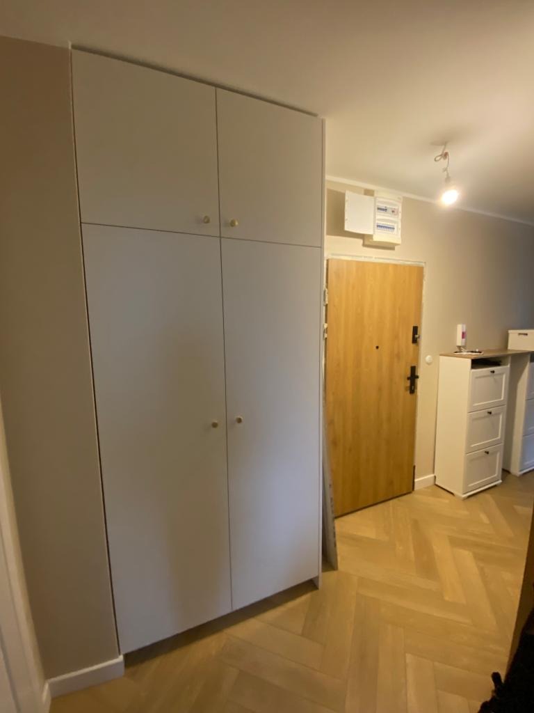 Wysoka, jasnoszara szafa wnękowa z czterema drzwiami i okrągłymi, złotymi uchwytami, dopasowana do wysokości pomieszczenia, na tle parkietu ułożonego w jodełkę.