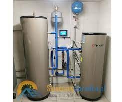 Dwa stojące zbiorniki buforowe z podłączeniami hydraulicznymi, widoczne rury i osprzęt w pomieszczeniu technicznym.