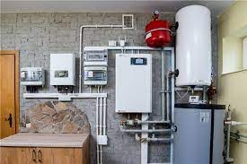 Instalacja grzewcza w pomieszczeniu technicznym: biały piec, szary zbiornik akumulacyjny, czerwony zbiornik wyrównawczy, rozdzielnica elektryczna i liczniki na ścianie z imitacją kamienia.