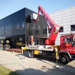 Czerwony podnośnik koszowy AlpiTech z pracownikami w koszu przy czarnym, przeszklonym biurowcu, widoczne podpory stabilizujące, kostka brukowa i trawnik.