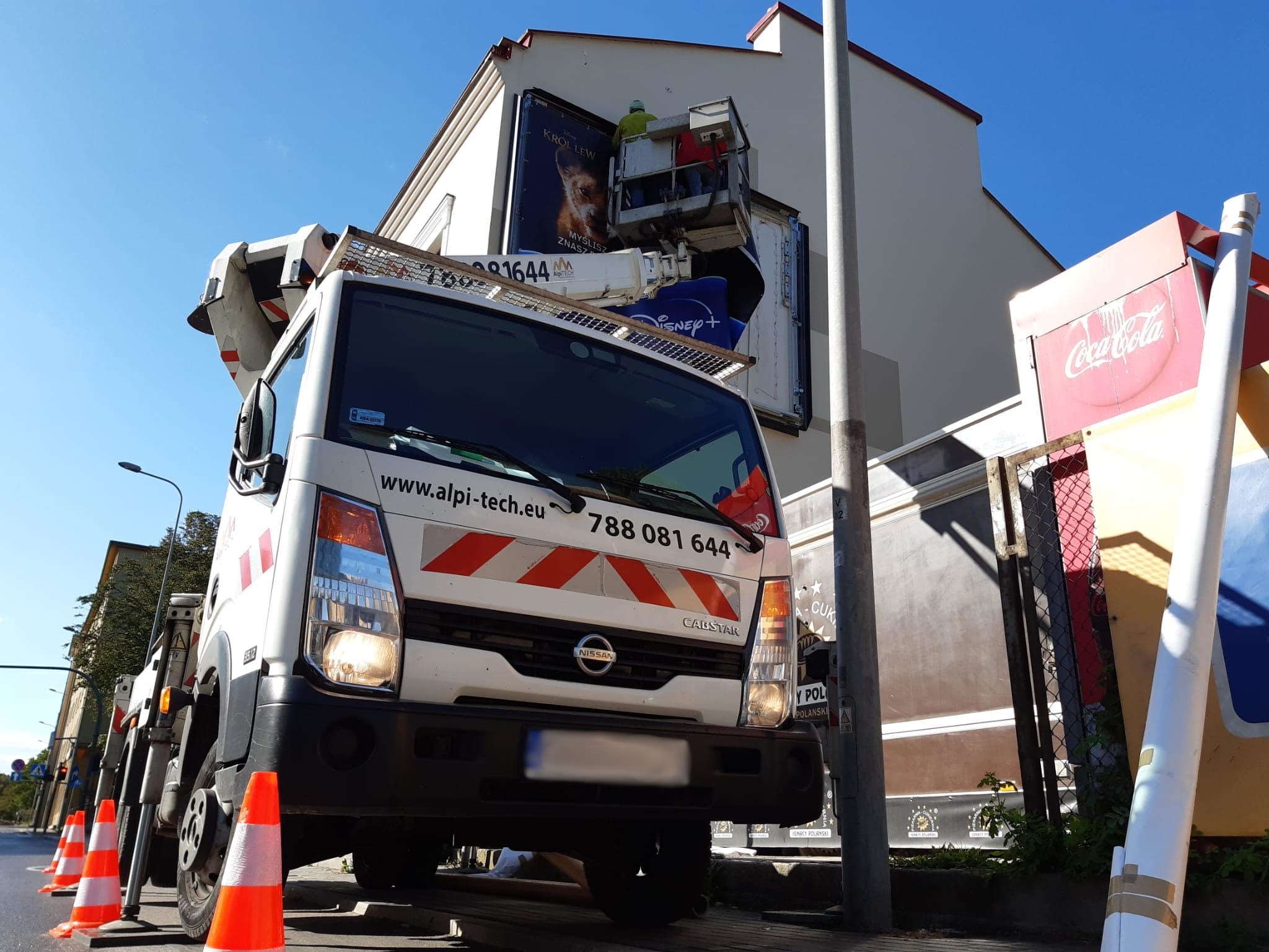 Pracownicy na podnośniku koszowym montują duży plakat filmowy 'Król Lew' na ścianie budynku, widoczny samochód firmy Alpi-tech z numerem telefonu i zaparkowane pachołki drogowe.