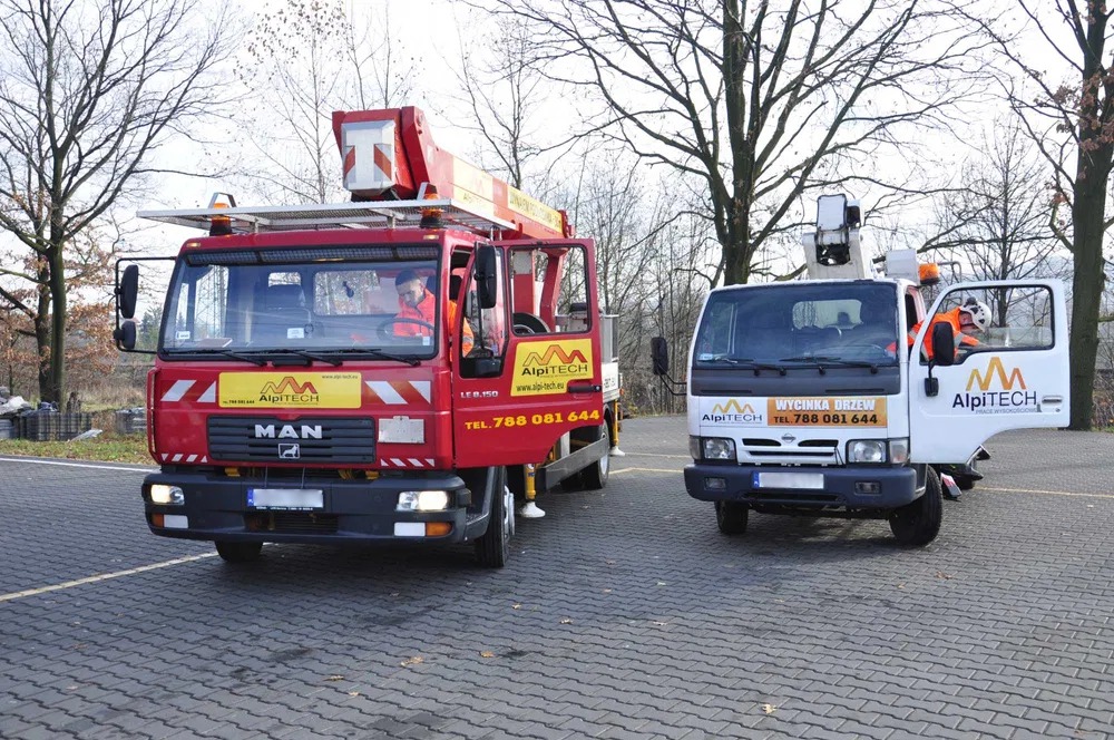Dwa samochody z podnośnikami koszowymi firmy AlpiTECH, jeden czerwony MAN, drugi biały Nissan, oba z otwartymi drzwiami, stojące na brukowanym parkingu w pochmurny dzień.