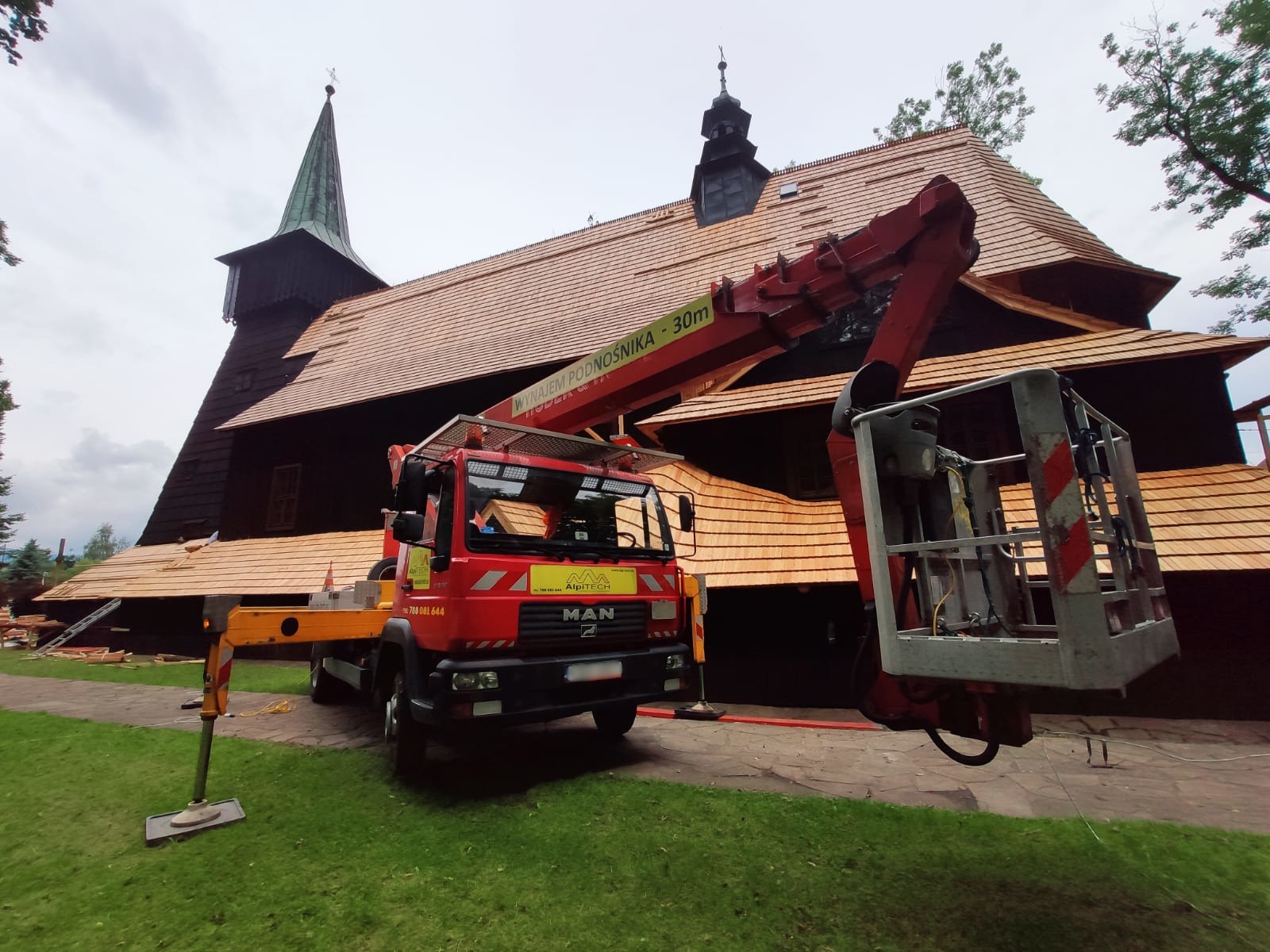 Czerwony podnośnik koszowy MAN na tle drewnianego kościoła z nowym, jasnym dachem z gontu, widoczne podpory stabilizujące pojazd na trawniku.
