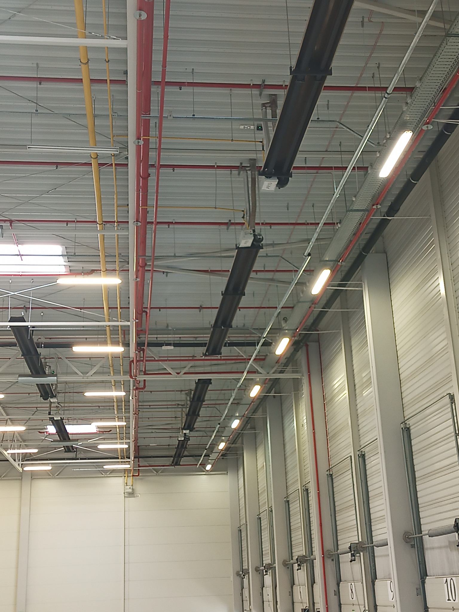 Wnętrze hali magazynowej z widoczną instalacją oświetleniową, rurami wentylacyjnymi w kolorach żółtym i czerwonym oraz systemem klimatyzacji pod sufitem, z widocznymi bramami segmentowymi w tle.