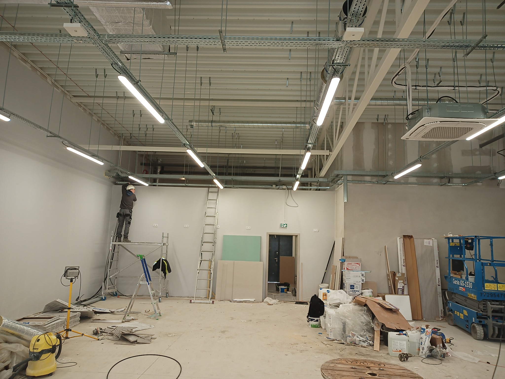 Pracownik w kasku na rusztowaniu montuje instalację pod sufitem w hali, widoczne kable, rury wentylacyjne, oświetlenie liniowe, klimatyzator kasetonowy, drabina, podnośnik koszowy oraz materiały...