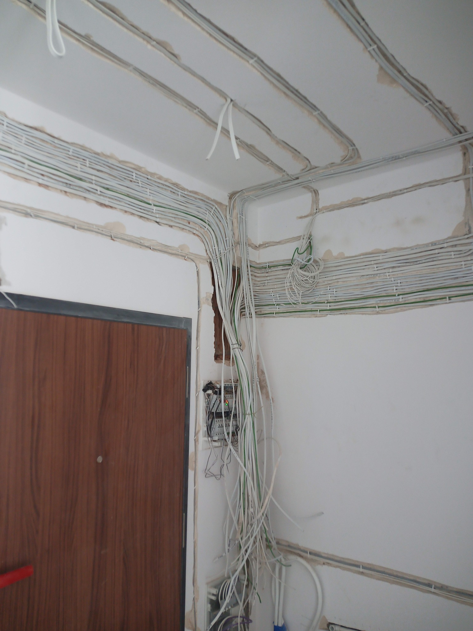Świeżo poprowadzone, białe kable elektryczne w bruzdach na ścianie i suficie w rogu pokoju, z widoczną puszką instalacyjną i fragmentem drewnianych drzwi.