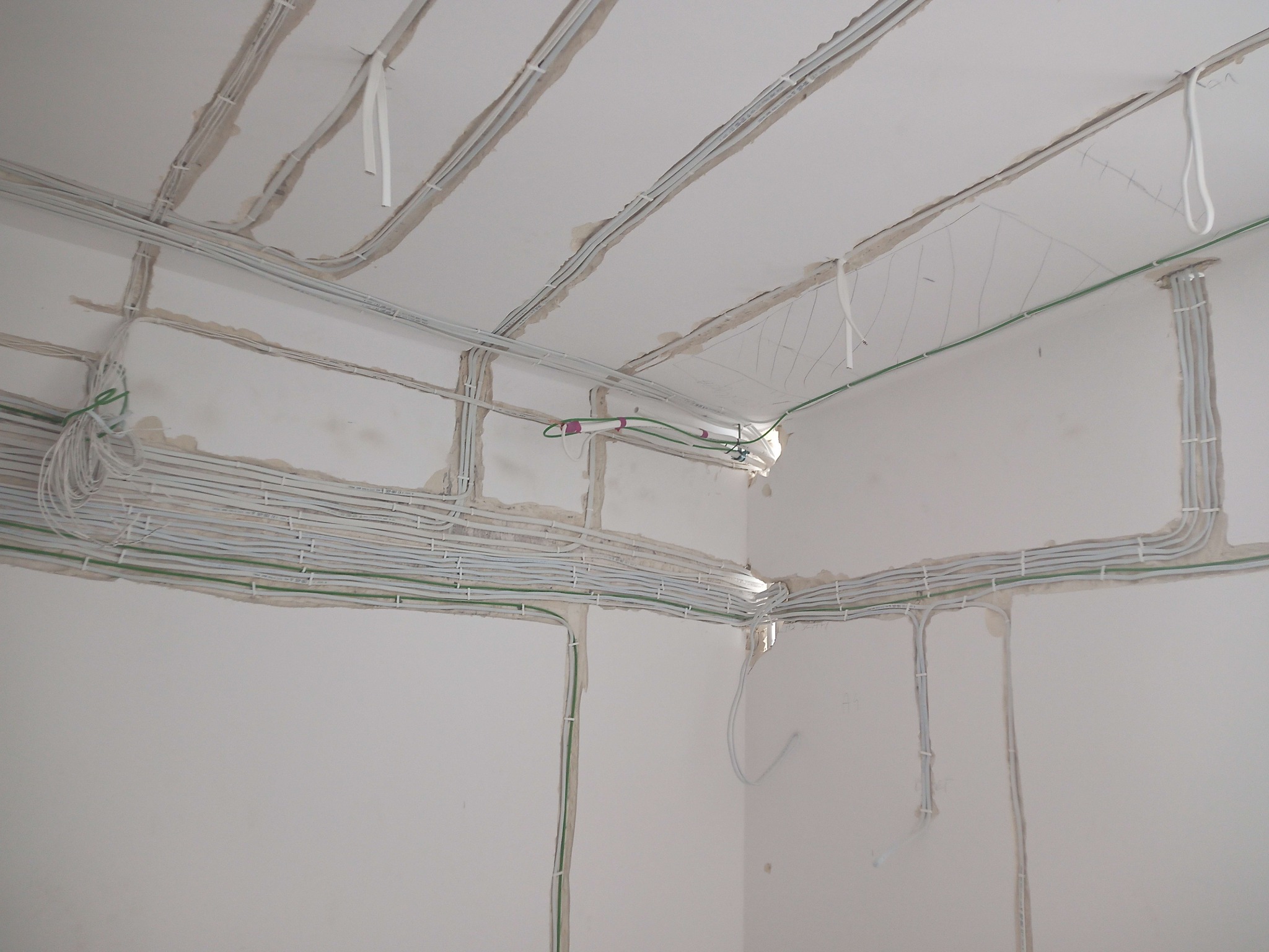 Świeżo położone przewody elektryczne w bruzdach na ścianie i suficie, przygotowane do tynkowania, z widocznymi zielonymi przewodami uziemiającymi i białymi uchwytami mocującymi.